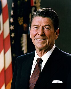 White House portrait of Reagan smiling in front of the U.S. and U.S. president flags, wearing a dark blue suit jacket with a white shirt and burgundy necktie.