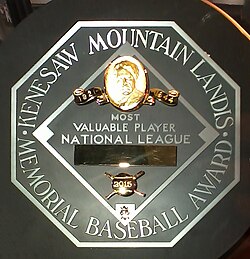 A black circle with an octagonal read "KENESAW MOUNTAIN LANDIS MEMORIAL BASEBALL AWARD". In the middle of the octagon is a baseball diamond which contains, from the top, Judge Landis' face in gold, "Most Valuable Player", the winner's league, his name in a gold rectangle, and his team.