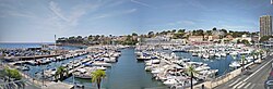 A panoramic view of Carry-le-Rouet harbour in France, showing rows of docked boats and yachts surrounded by seaside buildings and palm-lined streets under a clear sky.